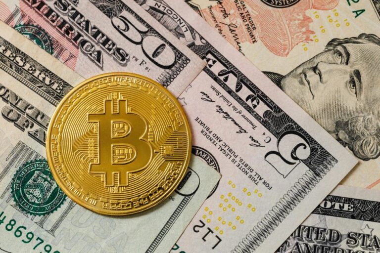 Auksinė „Bitcoin“ moneta padėta ant JAV dolerių banknotų.