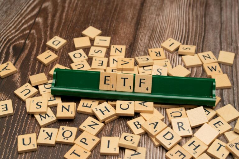 Medinės raidelių kaladėlės ant medinio paviršiaus, centre sudėliotas užrašas „ETF“, simbolizuojantis investavimą į biržoje prekiaujamus fondus.