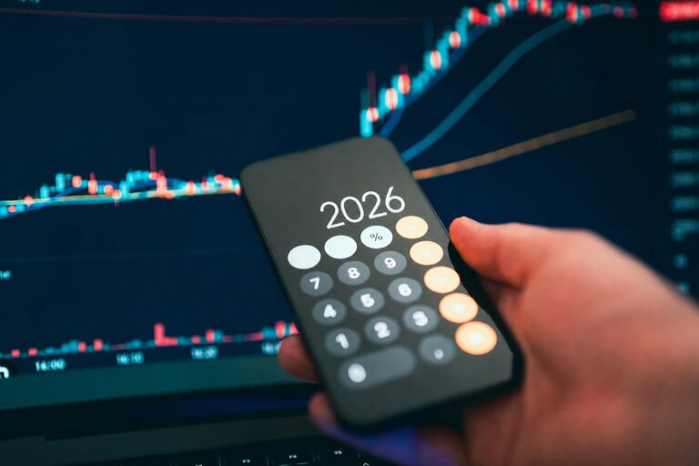 Rankoje laikomas telefonas su skaičiuotuvo ekrane matomu „2026“, fone – akcijų rinkos kainų grafikas, simbolizuojantis investavimo planavimą ir rinkos prognozes.